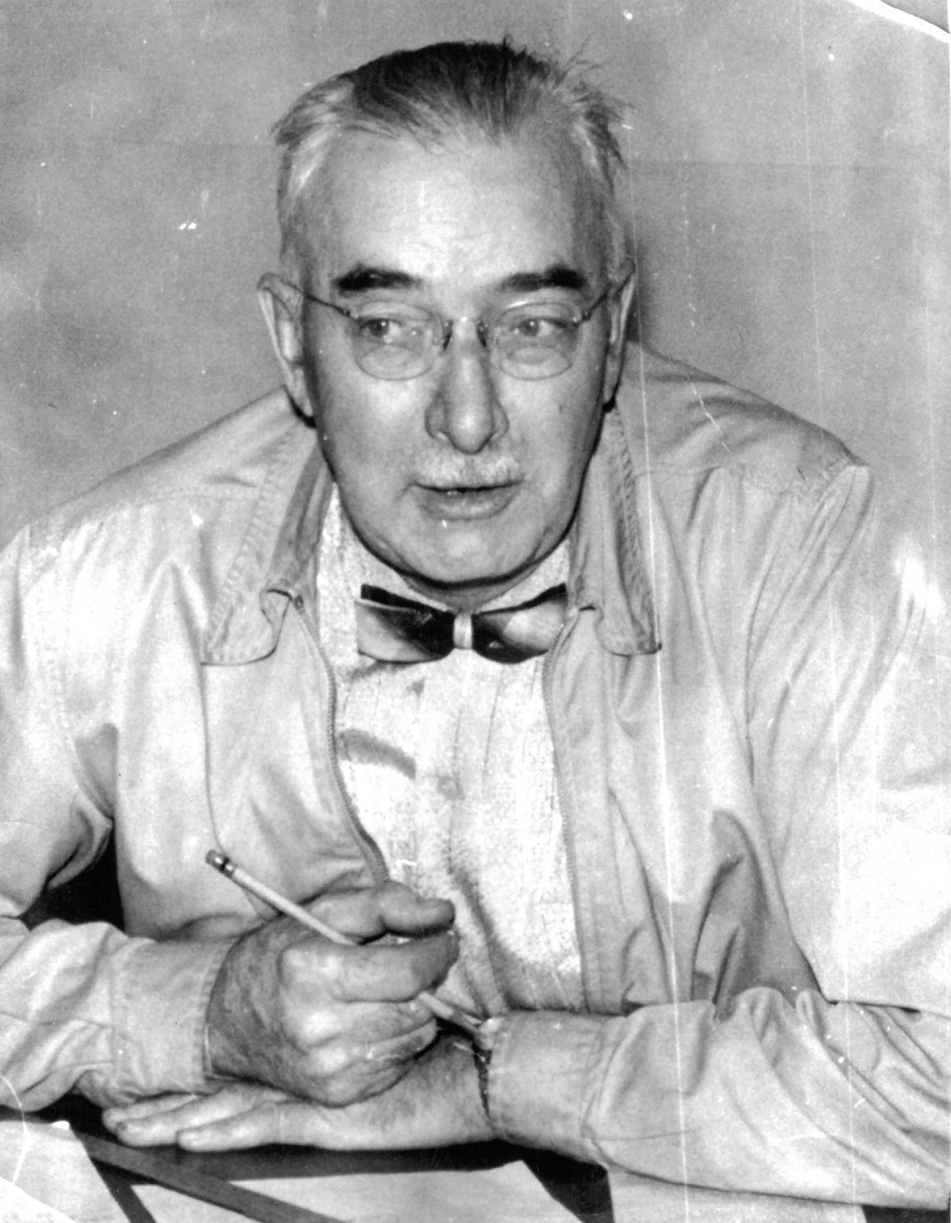 Neil Matheson sitting and holding a pencil, wearing glasses and a bowtie